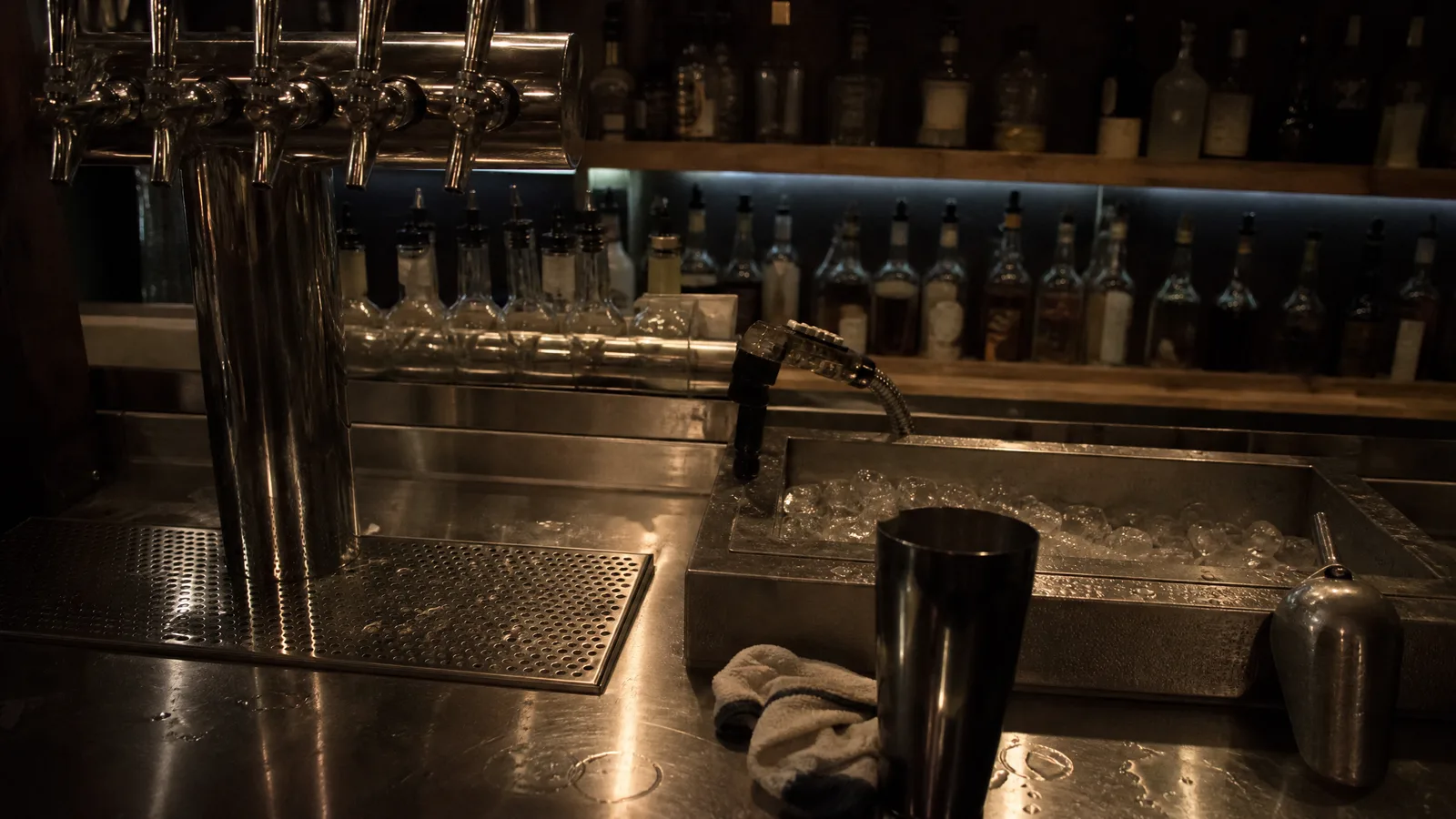 Bar back with a stainless tap tower, speed rail and shelves of unlabeled bottles under warm light